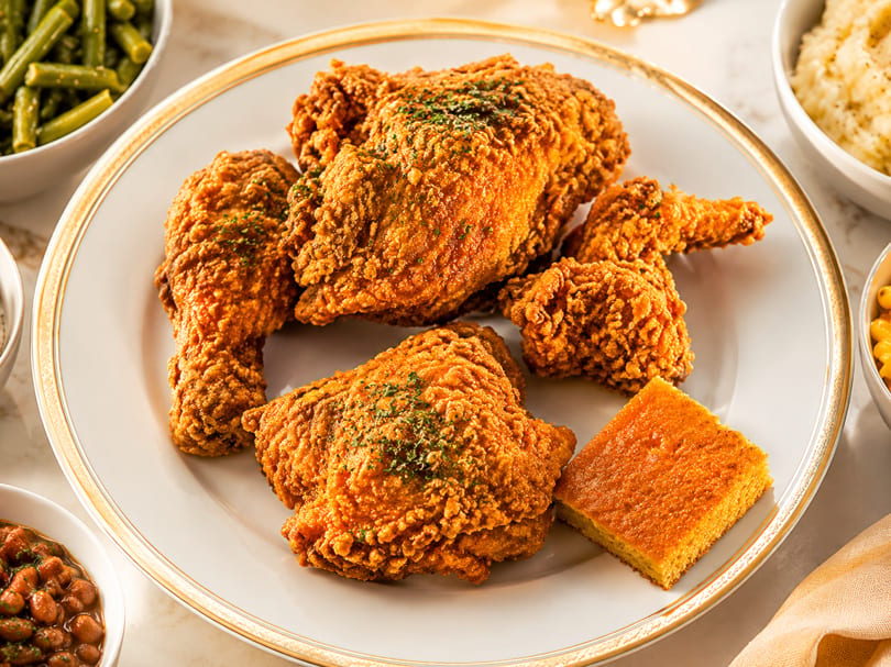 Golden fried chicken pieces and cornbread on a white plate, surrounded by side dishes.