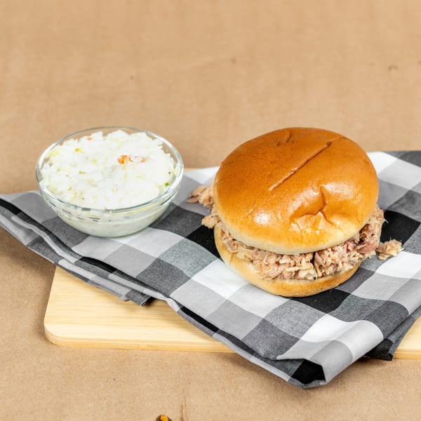Pulled pork sandwich on a bun with a side of coleslaw, on a checkered napkin and wooden board.