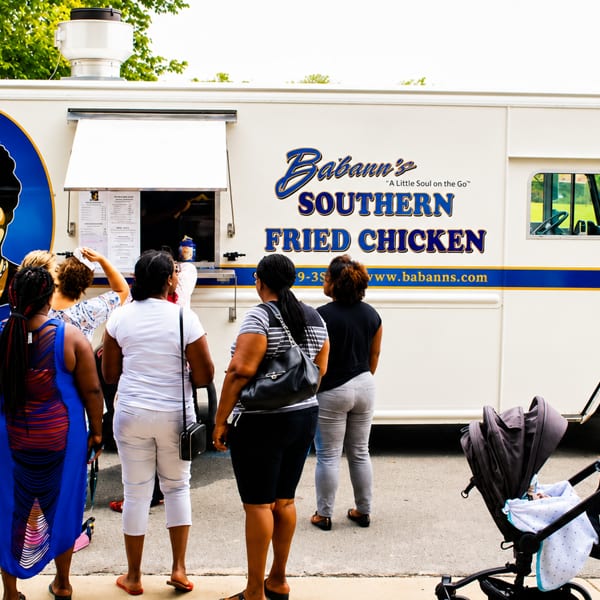 People queueing at a white Babann's Southern Fried Chicken food truck with a smiling woman's portrait.